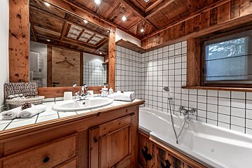 A rustic bathroom features a wood-paneled ceiling, vanity, and mirror, with white tiled walls, a sink, and a clean white bathtub.