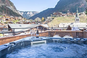 A bubbling hot tub with wine glasses on a wooden deck, overlooking a scenic alpine village surrounded by mountains.