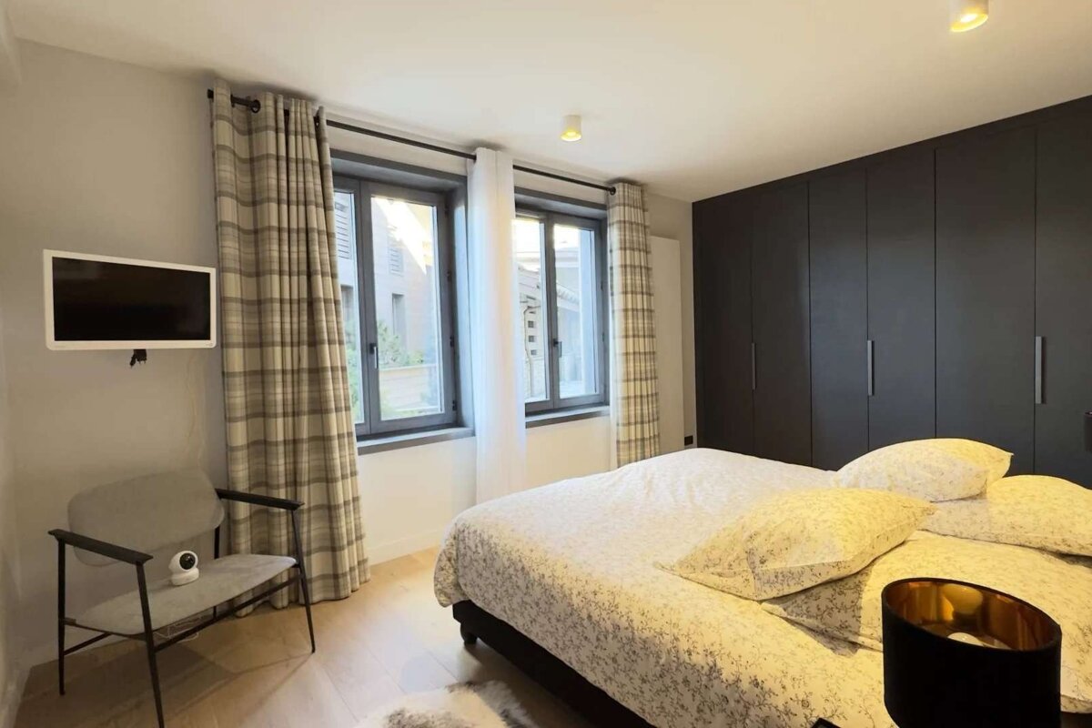 Modern bedroom with a patterned bed, dark built-in wardrobe, two windows with plaid curtains, wall-mounted TV, and an armchair with a security camera.