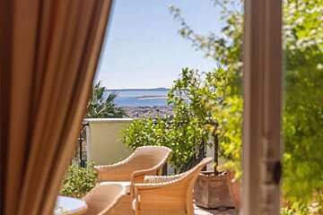A sunny balcony with wicker chairs and green plants overlooks a coastal city and a tranquil bay under a clear blue sky.