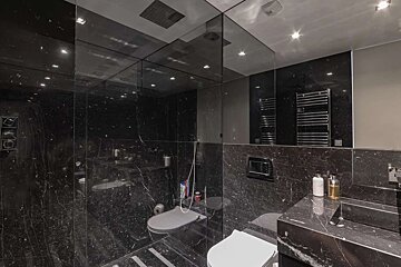 A luxurious, modern bathroom featuring dark, veined marble on all surfaces. Mirrored walls and bright spotlights create a dramatic, expansive feel.