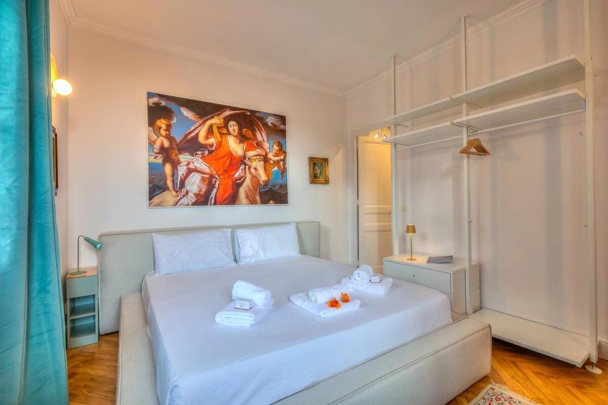 A bright bedroom featuring a white double bed under a large classical painting, with minimalist open wardrobe shelving on the right.