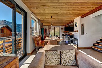 A spacious chalet living room with a wood-paneled ceiling, open dining area, modern kitchen, and large windows offering picturesque mountain and cabin views.