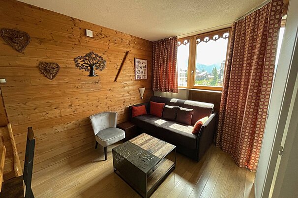 Cozy, chalet-style living room with wood-paneled walls, a dark sofa, gray armchair, unique coffee table, and sunlit window overlooking mountain views.