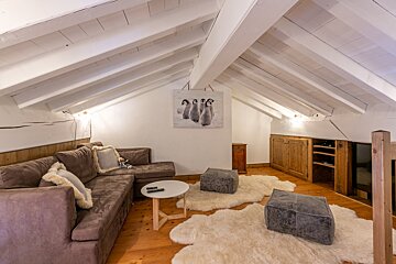 Cozy attic living room with a brown L-shaped sofa, white sloped beamed ceiling, wooden floors, furry rugs, and a penguin picture.