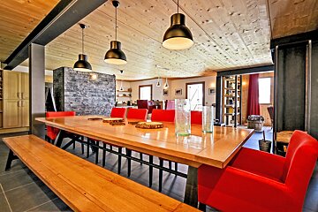 Modern rustic dining room with a long wooden table, bright red chairs, wooden bench, black pendant lights, and a wood-paneled ceiling.