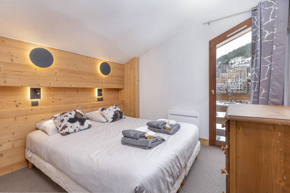 Cozy chalet bedroom featuring a large bed with cow-print pillows, wood wall paneling, and a balcony overlooking a snowy alpine village.