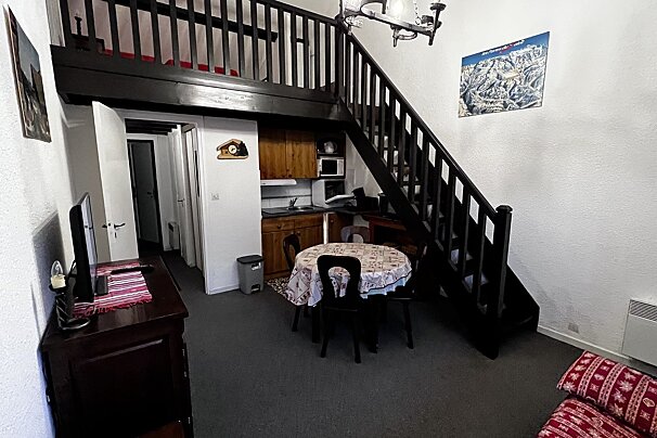 Small rustic apartment featuring a dining area, a kitchenette under a mezzanine loft, and a dark wooden staircase.