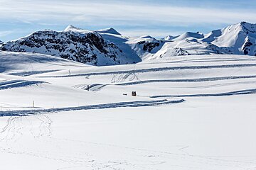 Vast, bright snowy mountain landscape with ski tracks and a small brown hut under a blue sky, surrounded by rugged peaks.