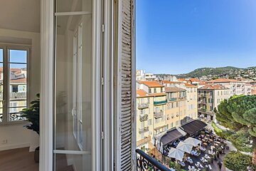 View from a balcony overlooking a charming European town with colorful buildings, red roofs, a lively outdoor restaurant, and distant green hills.