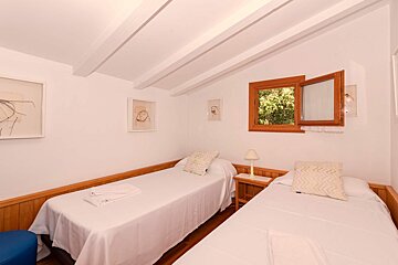 A bright twin bedroom with white walls, wooden ceiling beams, and wainscoting. It features two single beds, a small bedside table, and an open window revealing greenery.
