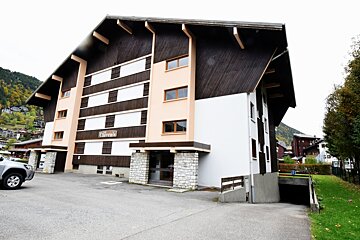 A multi-story brown wooden building with balconies is nestled in deep snow, surrounded by snow-capped mountains under a clear blue winter sky.