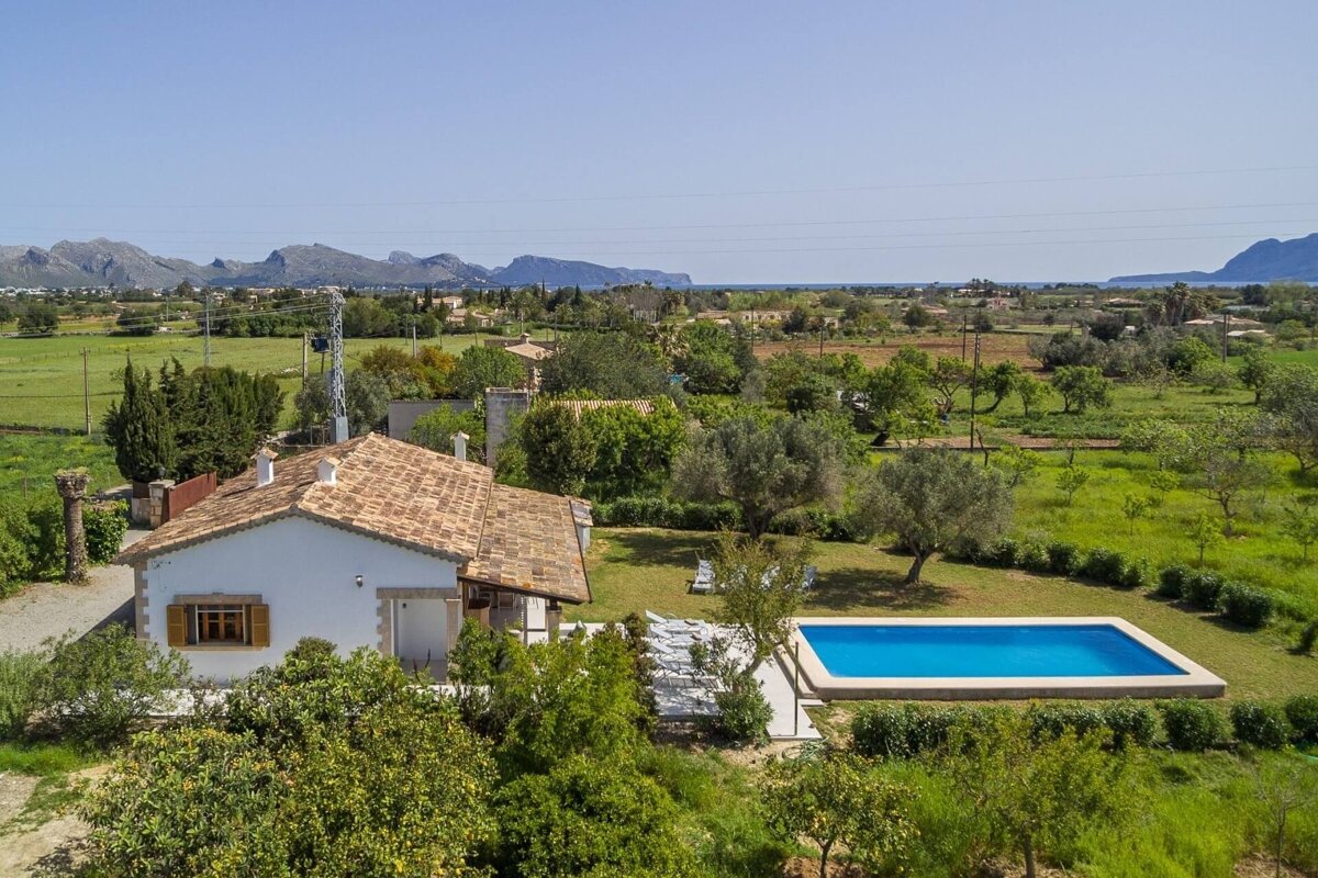 Aerial view of a white farmhouse with a blue pool, nestled in a vibrant green landscape with olive trees, distant mountains, and the sea visible under a clear sky.
