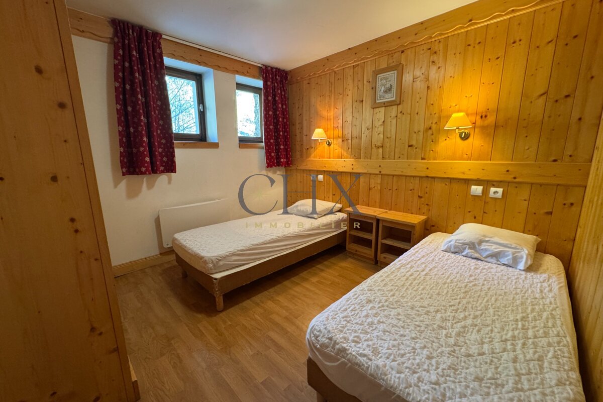 Cozy wood-paneled bedroom featuring two twin beds, rustic wooden walls, two windows with red curtains, and warm lighting.