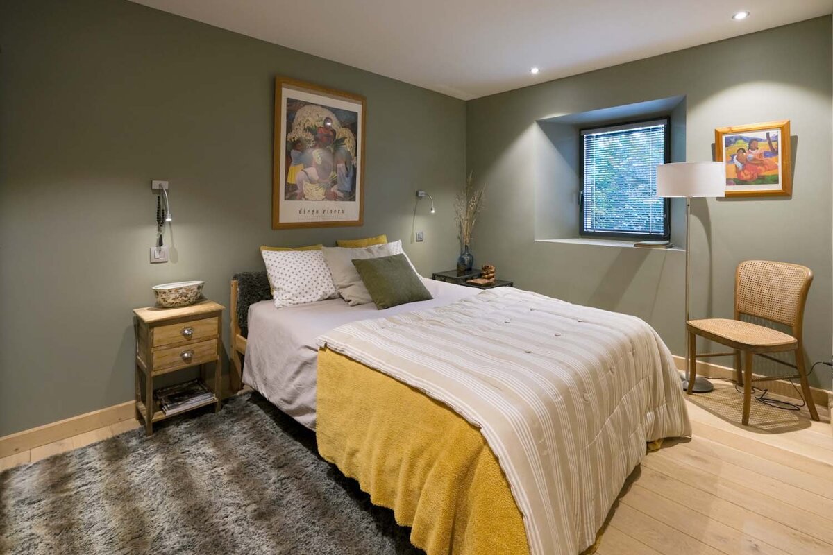 An inviting bedroom featuring olive-green walls, a bed with yellow and striped bedding, wooden furniture, and framed art, illuminated by natural light.