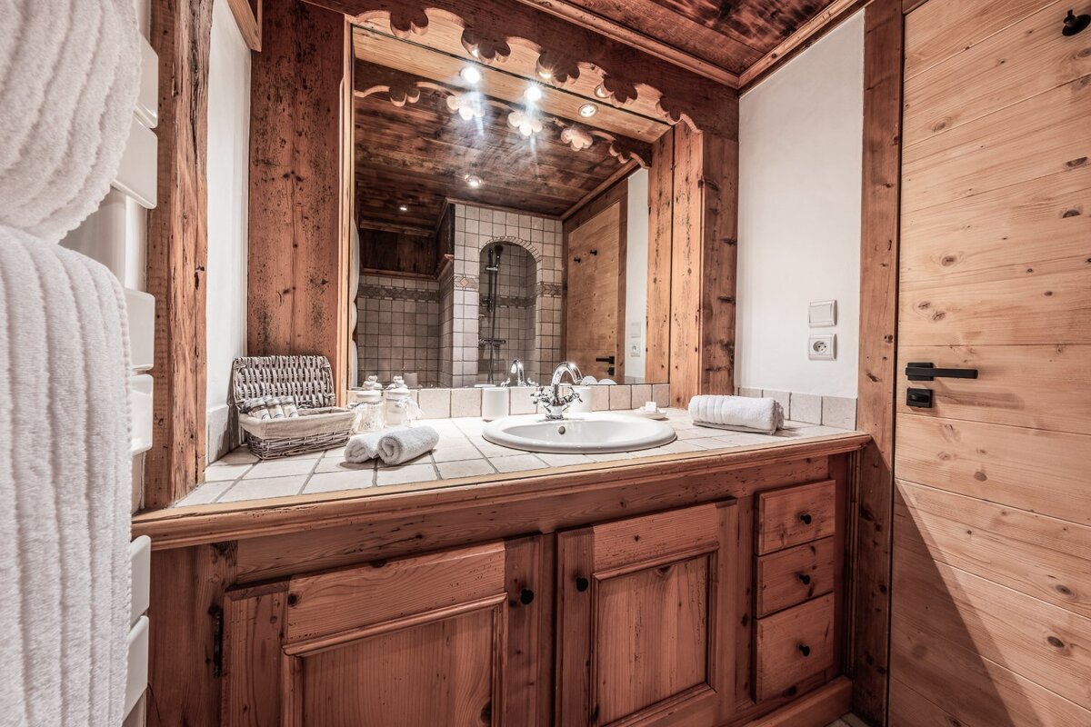 Rustic chalet-style bathroom featuring wooden cabinets, a large mirror, a tiled vanity with a white sink, and rolled towels.