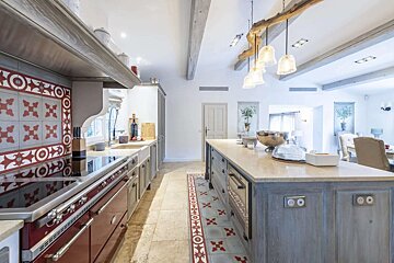 A spacious, rustic-chic kitchen with a red range, ornate tilework, a large island, exposed wooden beams, and a unique chandelier.