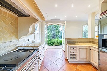 A bright kitchen with terracotta floor tiles, light cabinets, and granite counters opens to a lush garden through large French doors.