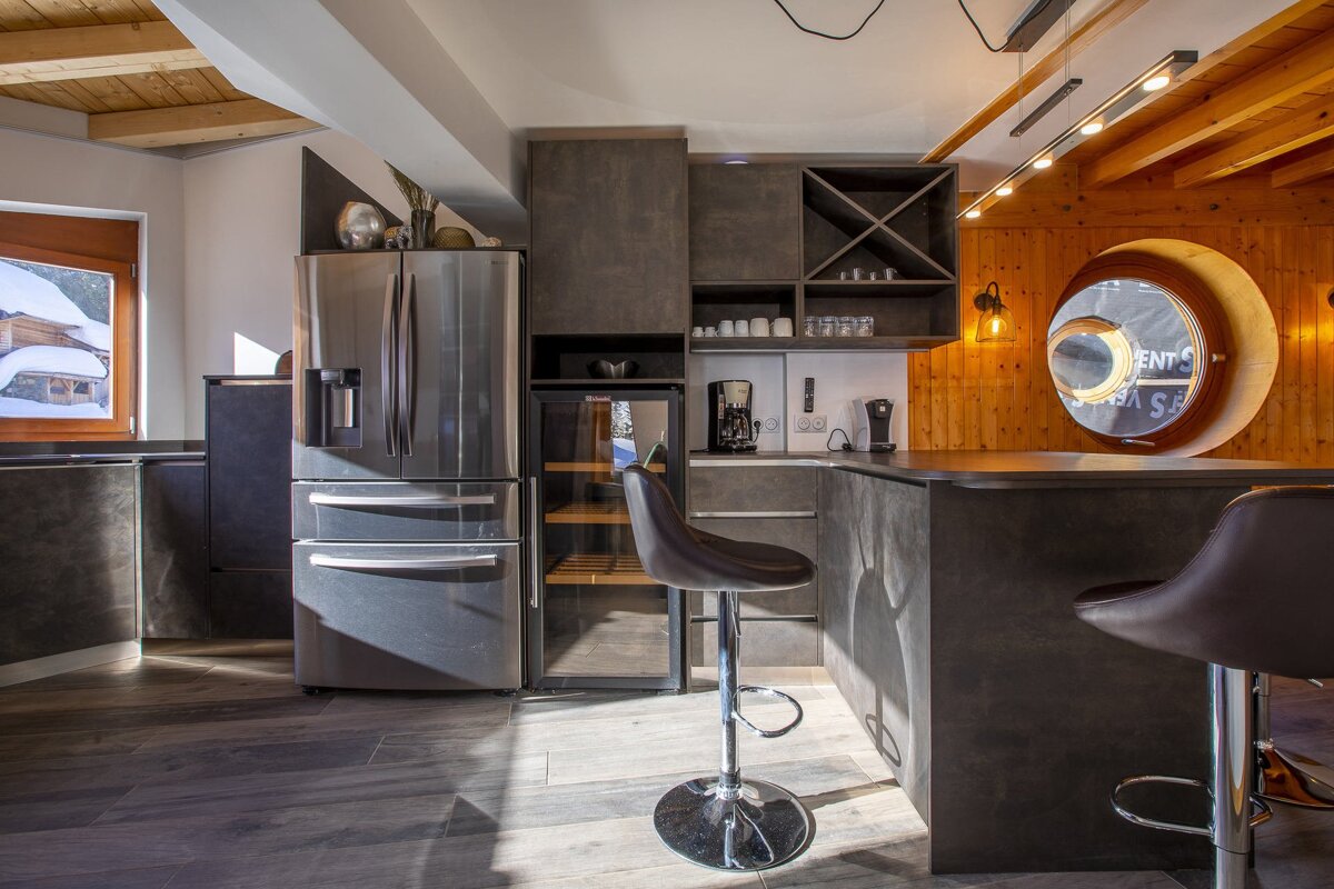 Modern kitchen with dark grey cabinets, stainless steel fridge, bar stools, and wood-paneled walls with a round window.