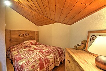 A cozy bedroom with a sloped wooden ceiling, a single bed with a red patchwork quilt, and a wooden headboard. A dresser holds a teddy bear and lamp.
