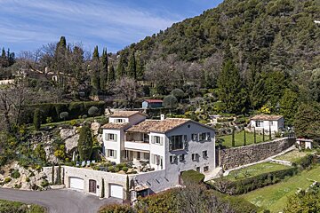 Aerial view of a large hillside house with terracotta roof and green shutters, surrounded by terraced gardens and backed by a dense forest.