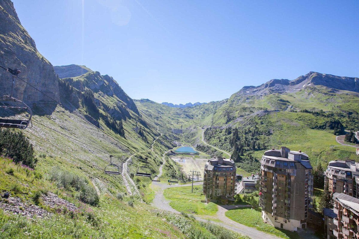 A verdant mountain valley featuring ski lifts, a clear blue lake, and several modern apartment buildings under a bright, sunny sky.