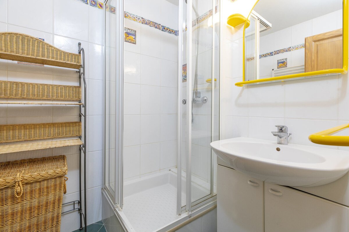 A small bathroom with white tiled walls, a shower cabin, sink, wicker storage shelves, and a yellow-framed mirror.