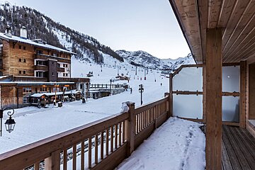 View from a wooden balcony overlooking a snowy ski resort. Charming timber buildings, ski slopes with lifts, and distant mountains fill the scene.