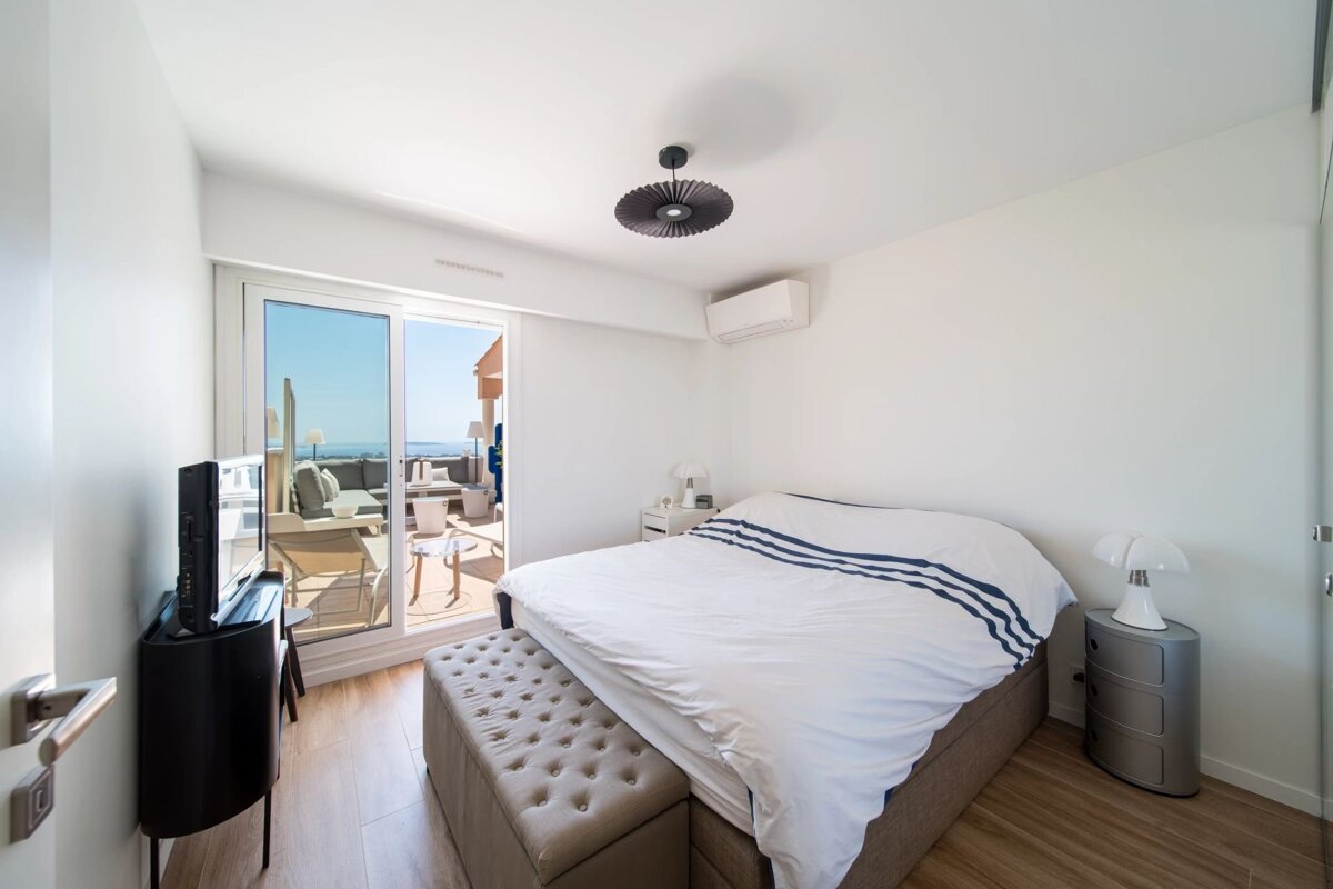 Bright, modern bedroom with white decor, a large bed, wooden floors, and a sliding glass door leading to a balcony with an ocean view.