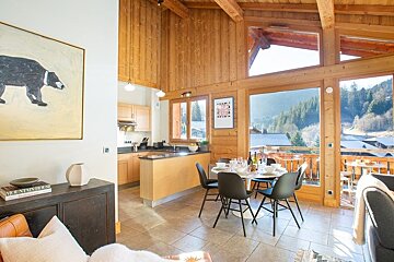 Cozy wooden chalet interior featuring a dining table, open kitchen, and large glass doors opening to scenic mountain views.