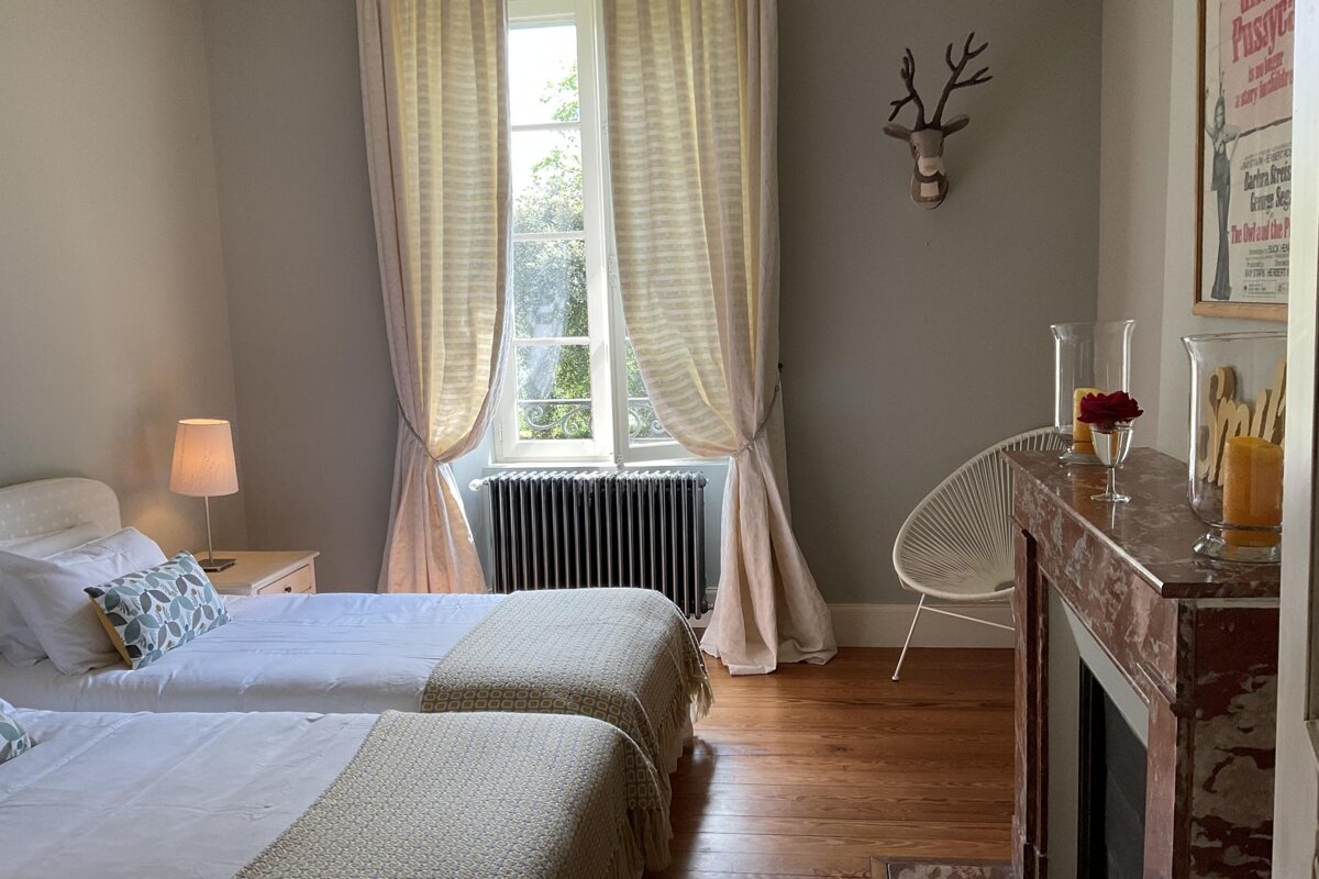 Cozy bedroom featuring two single beds, a large sunlit window with flowing curtains, a fireplace, and a decorative deer head.