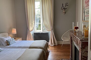 Cozy bedroom featuring two single beds, a large sunlit window with flowing curtains, a fireplace, and a decorative deer head.