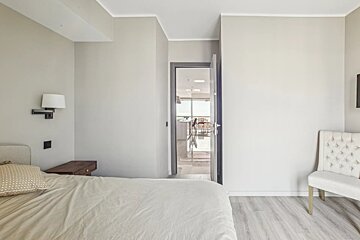 A bright, neutral bedroom features a partial view of a bed, a wall lamp, and an open doorway leading to a modern living area with light flooring.