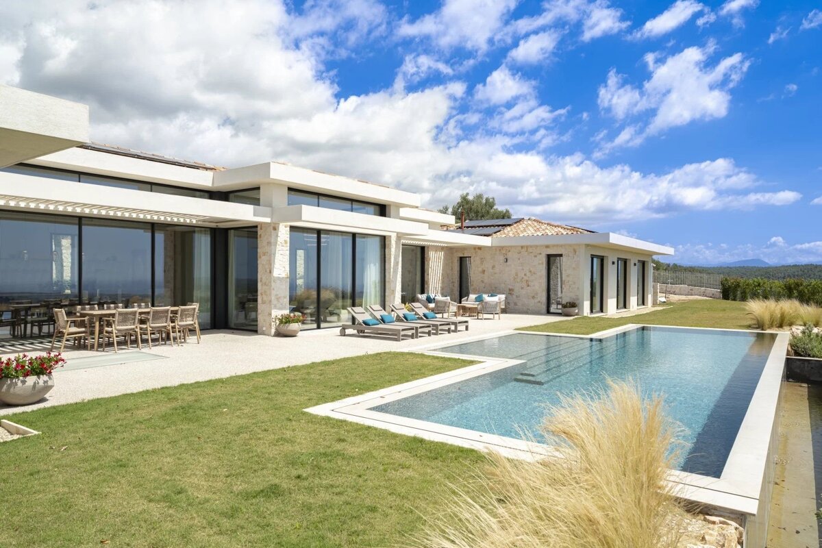 Modern stone villa with a sleek pool, green lawn, and patio under a bright blue sky, overlooking a distant landscape.