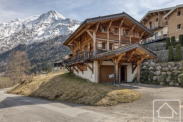 A rustic wooden chalet with balconies on a sunny hillside, backed by a majestic snow-capped mountain range and a forest.