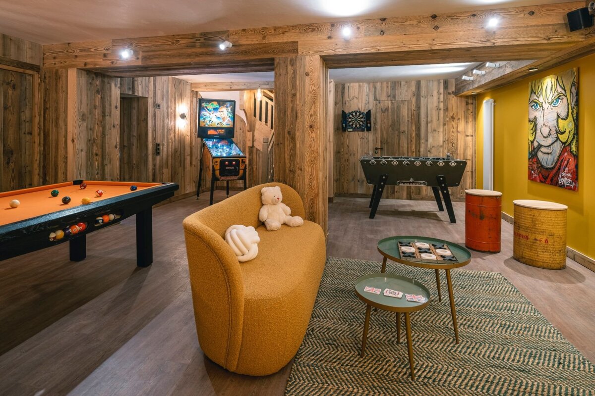 A rustic wood-paneled game room featuring a pool table, pinball, foosball, darts, a yellow sofa, and colorful artwork.