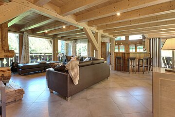 A rustic living room with exposed wooden beams, large windows, leather seating, and a cozy bar area, featuring natural wood and a bright, spacious feel.