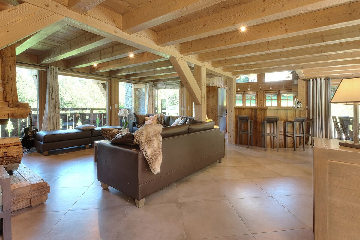 A rustic living room with exposed wooden beams, large windows, leather seating, and a cozy bar area, featuring natural wood and a bright, spacious feel.