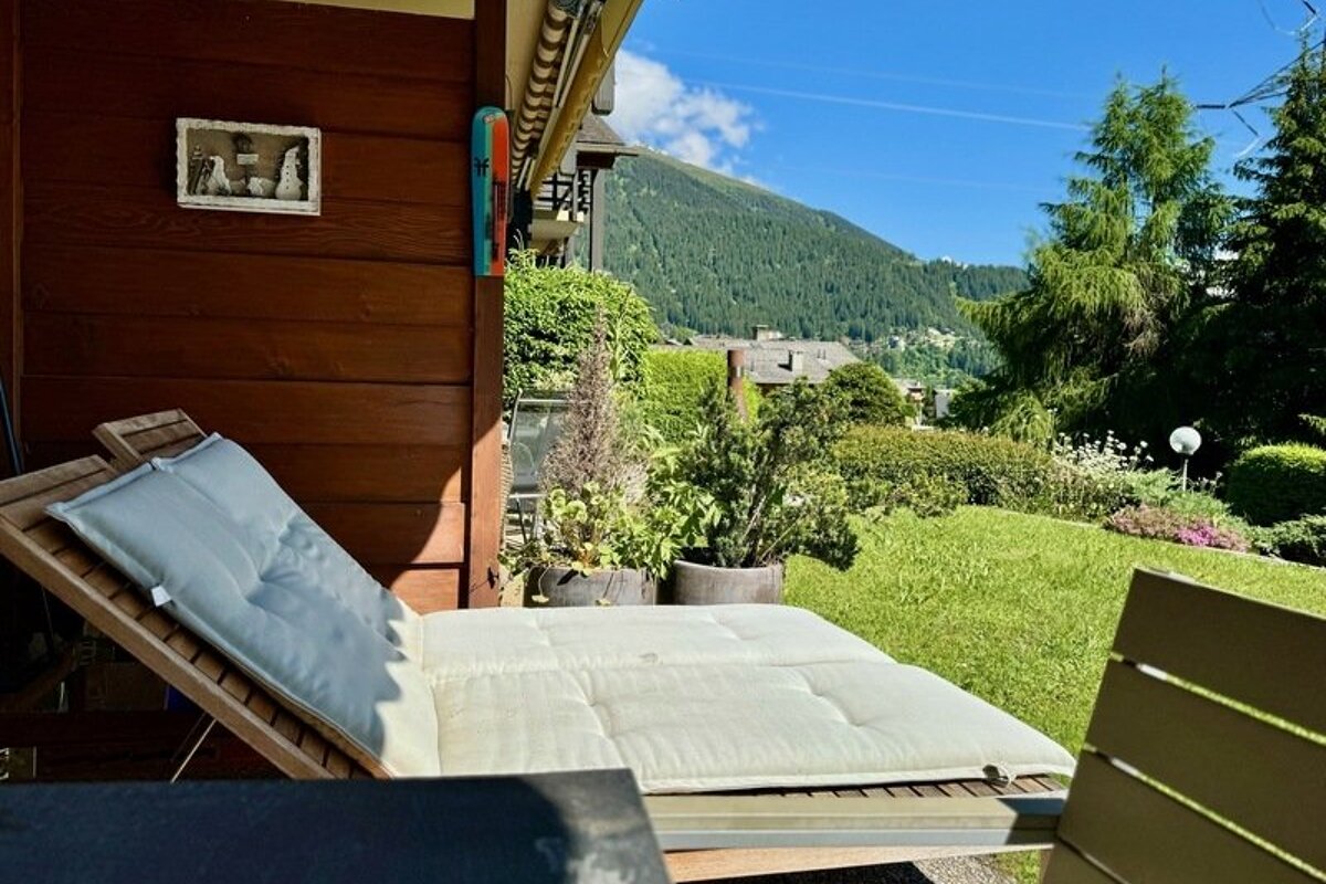 Wooden lounge chairs with white cushions on a sunny patio, overlooking a green garden and forested mountains.