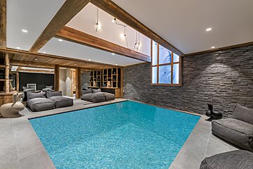 A luxurious indoor pool area with modern design, featuring wooden beams, a stone accent wall, cozy lounge seating, and a window overlooking a snowy mountain.