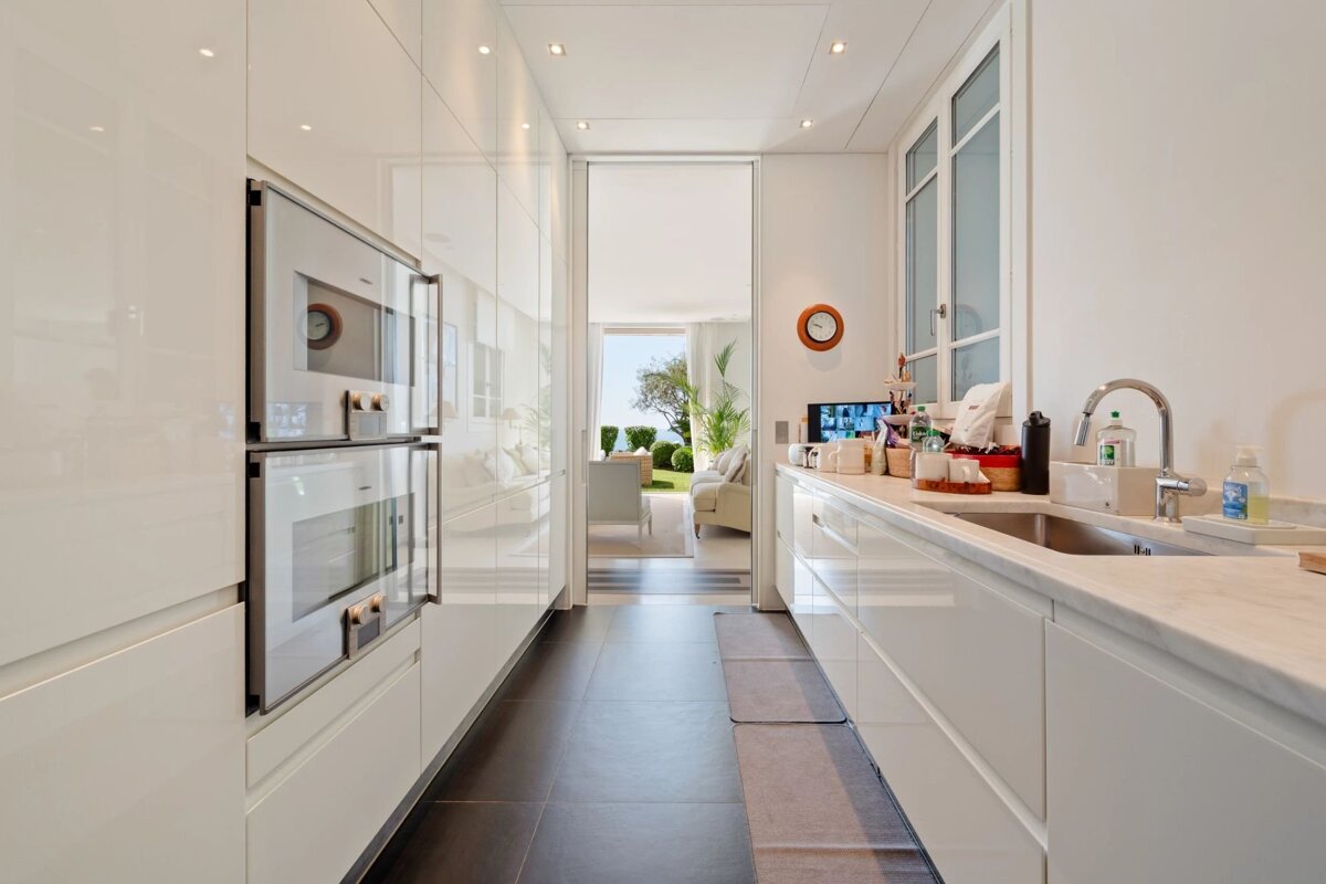 A sleek, modern white galley kitchen with built-in appliances, leading into a bright living room and outdoor patio.
