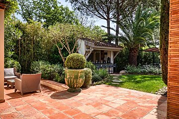An inviting outdoor patio with terracotta tiles, wicker furniture, and an outdoor bar, surrounded by lush green foliage and tall trees.
