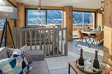 Stylish mountain chalet interior with a cozy living area, dining table, and large windows showcasing a snowy alpine landscape.