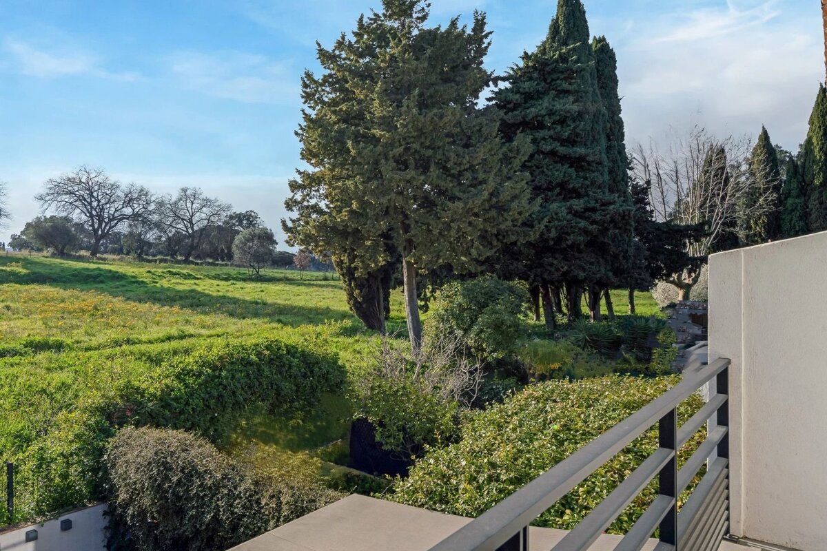 A balcony view overlooking a lush green field and tall cypress trees under a clear blue sky.