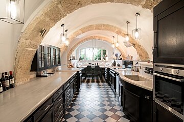 A long kitchen with stone arches, white vaulted ceilings, black cabinetry, and a checkerboard floor leading to a bright dining area with large windows.