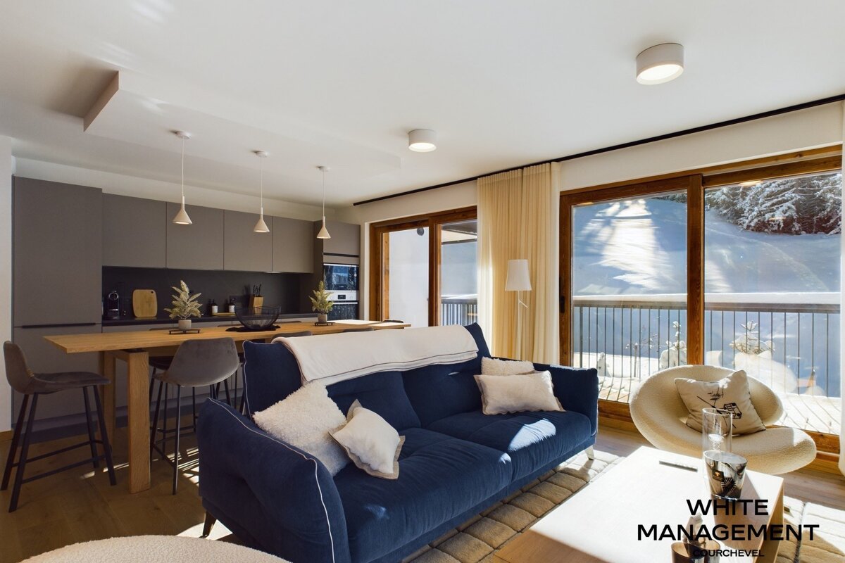 Modern open-plan living room and kitchen with a blue sofa, wooden island, and large windows revealing a snowy mountain view.