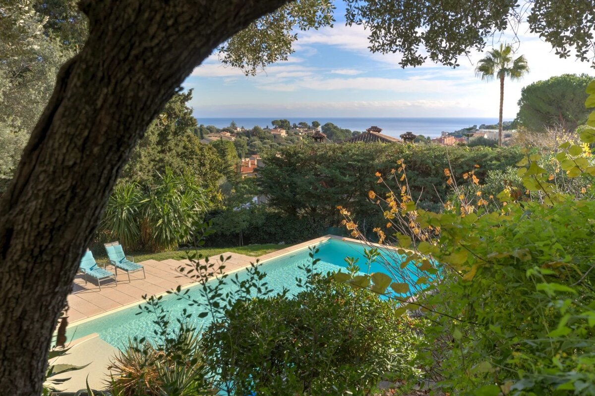 A beautiful view of a swimming pool framed by lush greenery, looking out over a coastal town towards the blue sea.