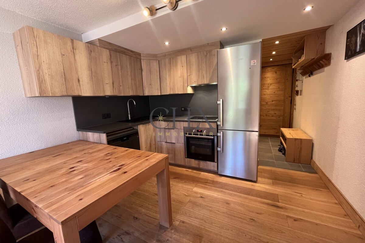 Modern wooden kitchen with light oak cabinets, a matching dining table, a stainless steel refrigerator, and sleek black countertops.