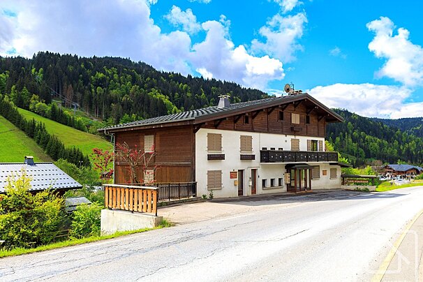 An alpine house with white and wood siding and a balcony stands beside a road, backed by green, forested mountains under a bright blue, cloudy sky.