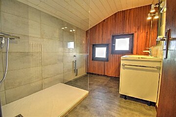 A modern bathroom features a walk-in glass shower, light tiled walls, and a warm wood-paneled section with a sink and two windows.